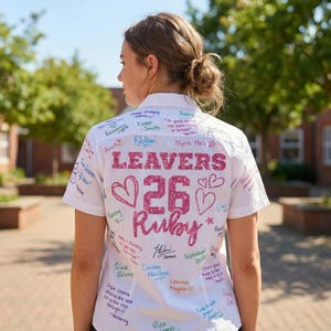 Peut inclure: Chemise blanche &agrave; manches courtes orn&eacute;e de messages manuscrits et de signatures. Le dos de la chemise affiche "LEAVERS 26 Ruby" en paillettes roses, avec des motifs de c&oelig;urs. Une personne porte la chemise.