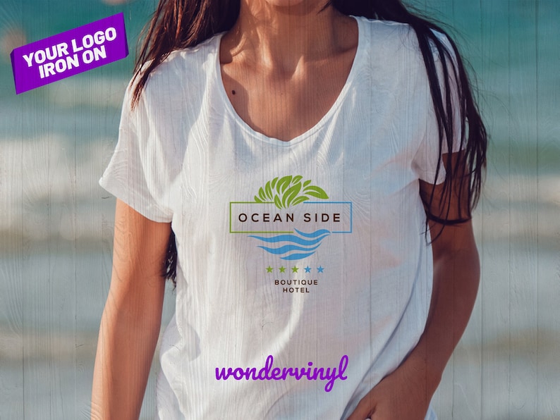 K&ouml;nnte beinhalten: Wei&szlig;es T-Shirt mit einem gr&uuml;nen, blauen und wei&szlig;en Logo, auf dem "Ocean Side Boutique Hotel" steht und ein Wellendesign. Der Text "wondervinyl" ist in Lila unter dem Logo gedruckt.