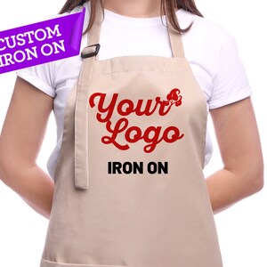 K&ouml;nnte beinhalten: Eine beige Sch&uuml;rze mit einem roten "Your Logo"-Design und dem aufgedruckten Text "Iron On". Die Sch&uuml;rze wird von einer Person getragen.