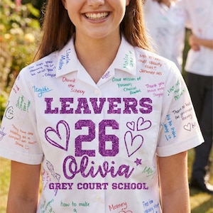Peut inclure: Une chemise blanche &agrave; manches courtes, couverte de messages manuscrits et de signatures. La chemise pr&eacute;sente le texte "LEAVERS 26 Olivia GREY COURT SCHOOL" en paillettes violettes. Des c&oelig;urs et des &eacute;toiles sont &eacute;galement dessin&eacute;s.