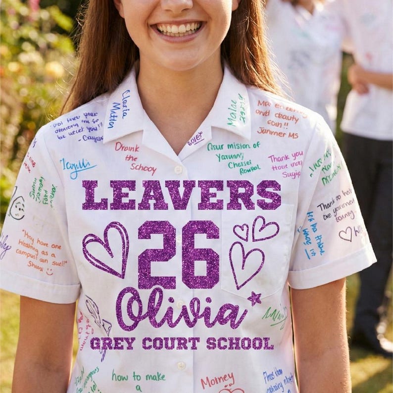 Peut inclure: Une chemise blanche &agrave; manches courtes, couverte de messages manuscrits et de signatures. La chemise pr&eacute;sente le texte "LEAVERS 26 Olivia GREY COURT SCHOOL" en paillettes violettes. Des c&oelig;urs et des &eacute;toiles sont &eacute;galement dessin&eacute;s.