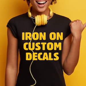 May include: A person wearing a black t-shirt with the text "IRON ON CUSTOM DECALS" in yellow lettering. The person is wearing yellow headphones around their neck.