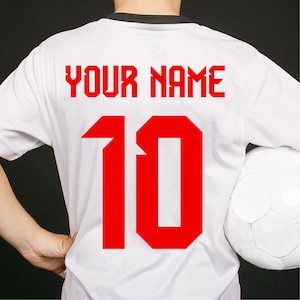 Op de afbeelding: Wit voetbal shirt met nummer 10 en de tekst "YOUR NAME" in rood gedrukt op de achterkant.