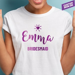 May include: White t-shirt with a purple iron-on design that says "Emma Bridesmaid" with a small heart and sun graphic above the name.