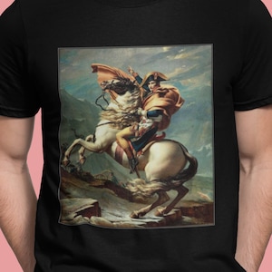 May include: A black t-shirt featuring a print of a painting depicting a man on horseback. The man is wearing a red and gold uniform and a white hat. The horse is white and is rearing up on its hind legs. The background is a landscape with mountains and clouds.
