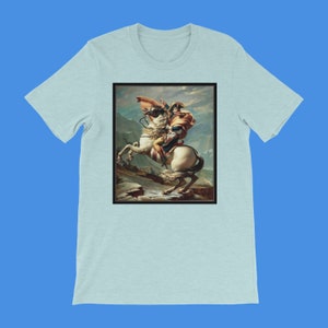 K&ouml;nnte beinhalten: Ein hellblaues T-Shirt mit einem Druck eines Gem&auml;ldes, das einen Mann auf einem Pferd zeigt. Der Mann tr&auml;gt eine blaue und rote Uniform und einen braunen Hut. Das Pferd ist wei&szlig; und b&auml;umt sich auf seinen Hinterbeinen auf. Der Hintergrund ist eine Berglandschaft.