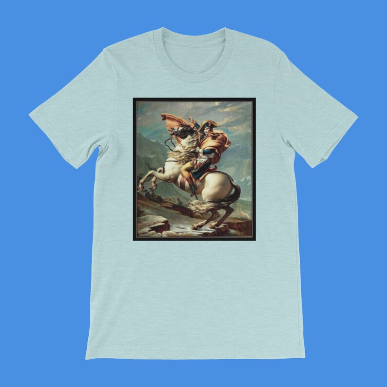 K&ouml;nnte beinhalten: Ein hellblaues T-Shirt mit einem Druck eines Gem&auml;ldes, das einen Mann auf einem Pferd zeigt. Der Mann tr&auml;gt eine blaue und rote Uniform und einen braunen Hut. Das Pferd ist wei&szlig; und b&auml;umt sich auf seinen Hinterbeinen auf. Der Hintergrund ist eine Berglandschaft.