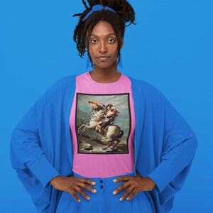 K&ouml;nnte beinhalten: Eine Frau tr&auml;gt ein pinkfarbenes T-Shirt mit einem Gem&auml;lde eines Mannes auf einem Pferd. Das Gem&auml;lde ist in einem schwarzen Rahmen. Die Frau tr&auml;gt einen blauen Cardigan und eine blaue Hose. Sie steht vor einem blauen Hintergrund.