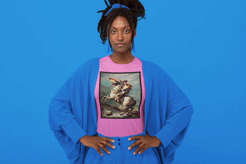 K&ouml;nnte beinhalten: Eine Frau tr&auml;gt ein pinkfarbenes T-Shirt mit einem Gem&auml;lde eines Mannes auf einem Pferd. Das Gem&auml;lde ist in einem schwarzen Rahmen. Die Frau tr&auml;gt einen blauen Cardigan und eine blaue Hose. Sie steht vor einem blauen Hintergrund.