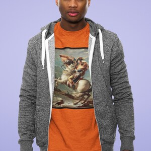 K&ouml;nnte beinhalten: Ein Mann tr&auml;gt einen grauen Rei&szlig;verschluss-Hoodie &uuml;ber einem orangefarbenen T-Shirt mit einem Gem&auml;lde eines Mannes auf einem Pferd. Das Gem&auml;lde ist in einem braunen Rahmen.