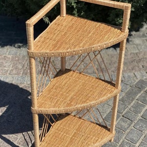 Puede incluir: Estantería esquinera de tres niveles hecha de mimbre tejido de color marrón claro. Los estantes son triangulares, con un patrón tejido. El marco está hecho de soportes verticales y tirantes diagonales. El estante superior tiene una pequeña barandilla.