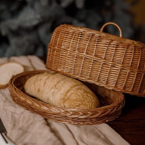 Op de afbeelding: Een goudbruin brood ligt in een ovale rieten mand met een deksel. Een sneetje brood en een gekarteld mes liggen op een beige doek. De mand is gemaakt van gevlochten riet.