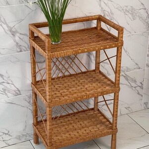 May include: A three-tiered, light brown wicker shelf unit. The shelves are rectangular and woven, with a decorative crisscross pattern on the sides. A clear glass vase with white flowers sits on the top shelf. The unit is freestanding.