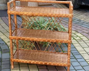 May include: A three-tiered wicker shelf unit with a natural brown finish. The shelf is made of woven wicker and has a diamond pattern on the sides. The shelf is standing on a brick patio.