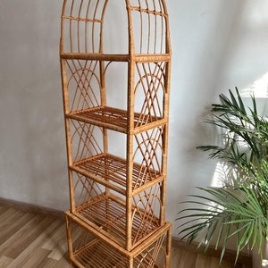 May include: A tall, arched, natural wicker shelf unit with four shelves. The shelf unit is made of woven rattan and has a natural brown color.