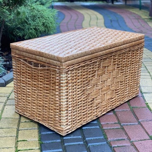 May include: A large, rectangular wicker storage basket with a lid. The basket is made of natural woven wicker and has a light brown color.