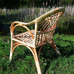 May include: A wicker chair with a woven back and seat, sitting in a grassy area near a pond. The chair is made of natural, light brown wicker.
