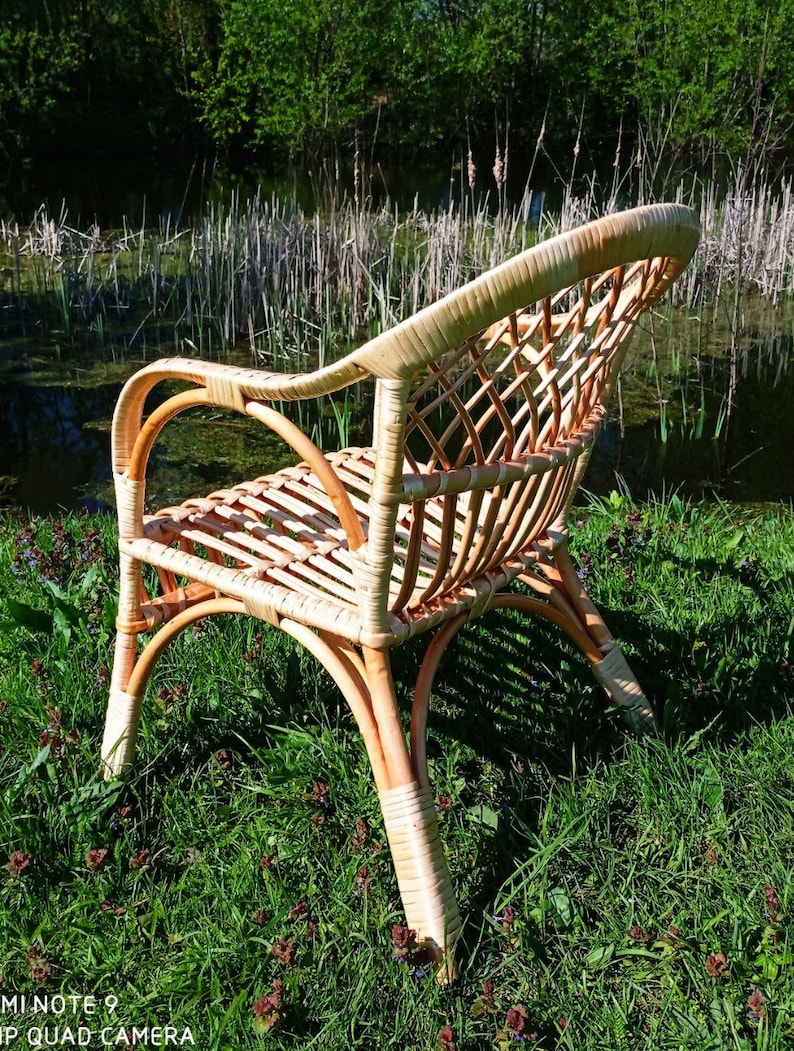 May include: A wicker chair with a woven back and seat, sitting in a grassy area near a pond. The chair is made of natural, light brown wicker.