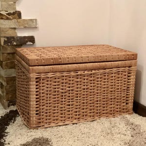 Puede incluir: Una cesta de almacenamiento de mimbre tejido con tapa. La cesta es de color marr&oacute;n claro y tiene una forma rectangular.