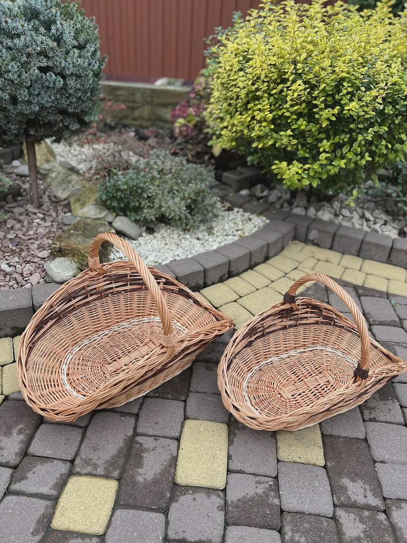 May include: Two woven wicker baskets with a natural light brown finish and a single arched handle. The baskets feature a distinctive asymmetrical, sloped shape with a flat base and a curved rim, showcasing a detailed interlaced willow pattern.