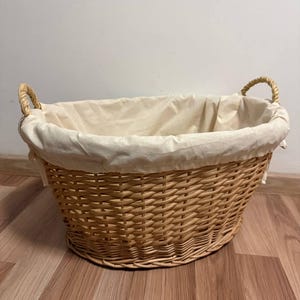 Puede incluir: Cesta de lavandería ovalada de mimbre tejido con un forro de tela color crema. La cesta tiene dos asas de cuerda y está sobre un suelo de madera. Un elemento práctico para el hogar.