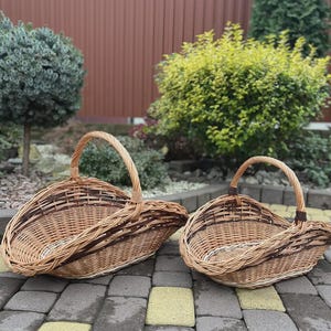 May include: Two oval-shaped woven wicker baskets with arched handles. The baskets feature a natural light brown finish with darker brown woven accents along the rims and sides. The construction consists of interlaced willow branches.
