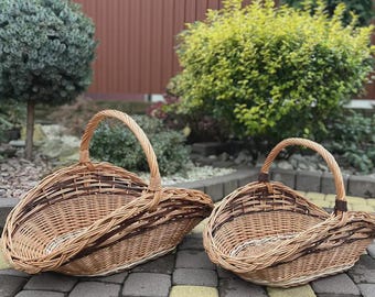 May include: Two oval-shaped woven wicker baskets with arched handles. The baskets feature a natural light brown finish with darker brown woven accents along the rims and sides. The construction consists of interlaced willow branches.