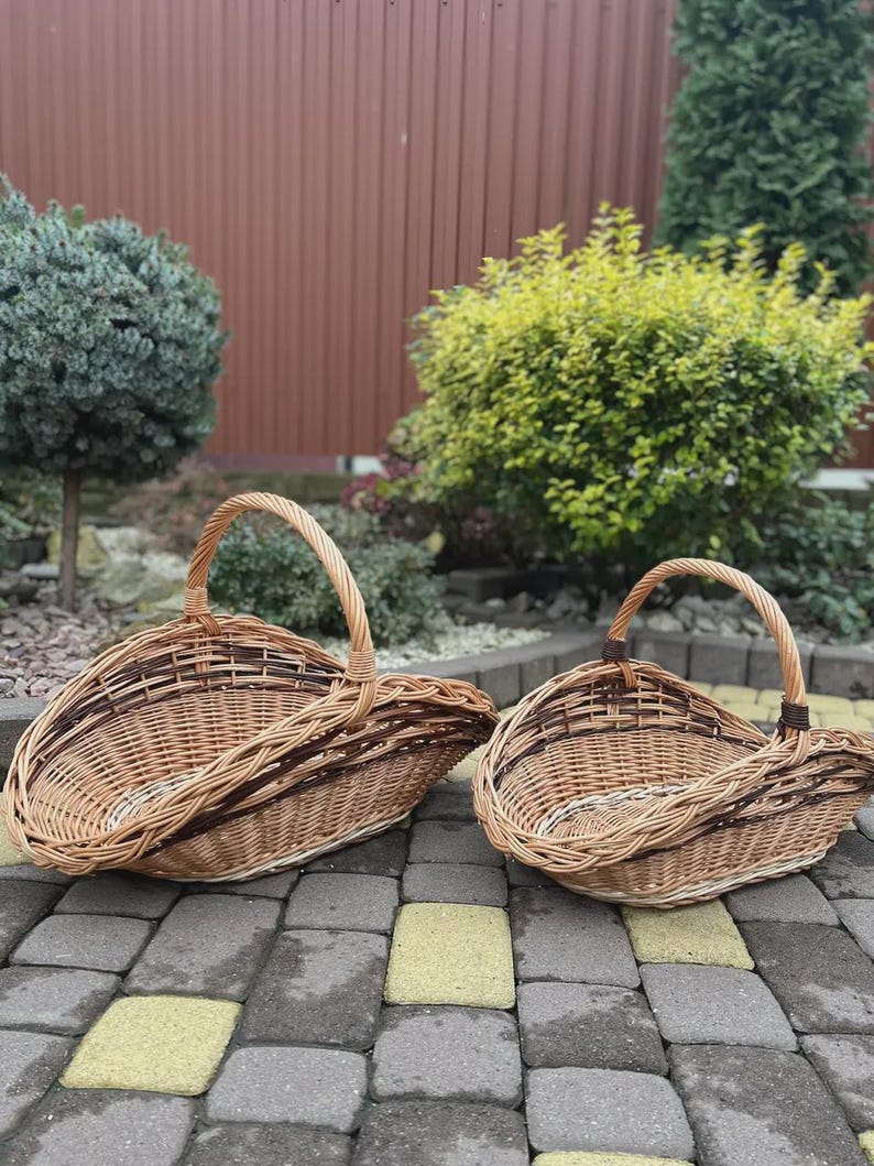 May include: Two oval-shaped woven wicker baskets with arched handles. The baskets feature a natural light brown finish with darker brown woven accents along the rims and sides. The construction consists of interlaced willow branches.
