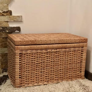 Puede incluir: Una cesta de almacenamiento de mimbre tejido natural con tapa. La cesta es de color marr&oacute;n claro y tiene una forma rectangular.
