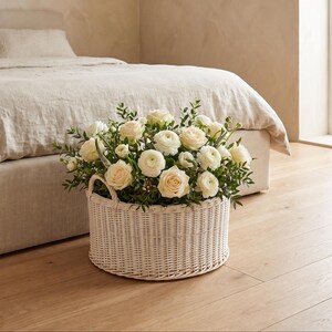 Pode incluir: Uma cesta redonda de vime trançado branco com duas alças laterais, preenchida com um arranjo denso de rosas e ranúnculos na cor creme, intercalados com pequenas folhas verdes e caules finos.