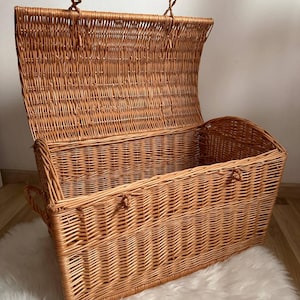 May include: A large, open wicker chest with a hinged lid. The chest is made of woven light brown wicker and has a rectangular shape with rounded edges. It features handles on the sides and a latch to secure the lid. The chest is sitting on a white fluffy rug.