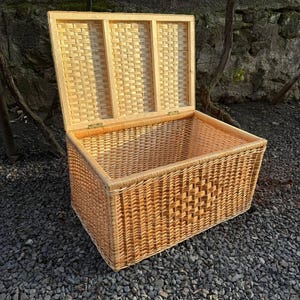 May include: A rectangular wicker storage chest with an open lid. The chest is made of woven light brown wicker and has a rectangular shape. The lid is also made of wicker and is attached to the chest with brass hinges. The chest is sitting on a bed of small gray stones.