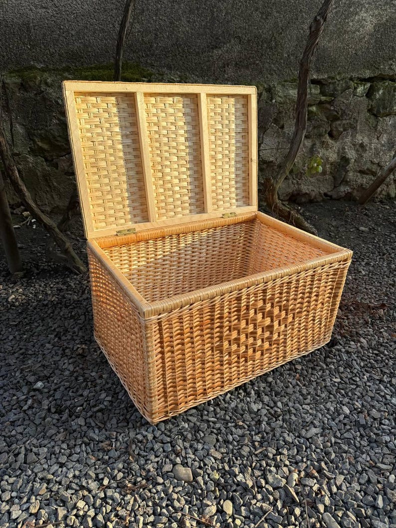 May include: A rectangular wicker storage chest with an open lid. The chest is made of woven light brown wicker and has a rectangular shape. The lid is also made of wicker and is attached to the chest with brass hinges. The chest is sitting on a bed of small gray stones.