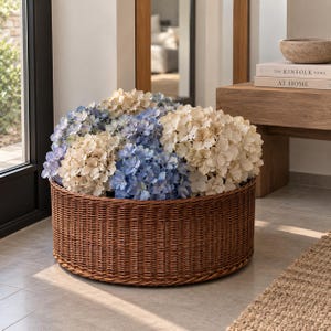 Könnte beinhalten: Ein großer, runder, brauner Korb aus geflochtenem Weidengeflecht, gefüllt mit einem dichten Arrangement aus getrockneten Hortensienblüten. Das Blumenarrangement zeigt eine Mischung aus zartblauen und cremeweißen Blütenblättern.