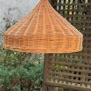 Pu&ograve; includere: Un paralume in vimini intrecciato di colore marrone chiaro, appeso a un soffitto in un ambiente esterno.