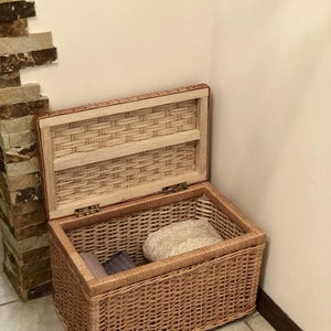 Puede incluir: Una cesta de almacenamiento de mimbre tejido con tapa, abierta para revelar tela doblada en el interior. La cesta es de color marr&oacute;n claro y tiene un acabado de madera natural.