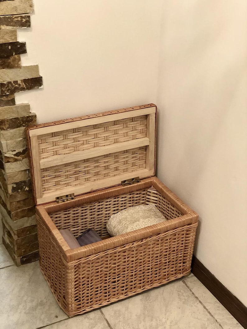 Puede incluir: Una cesta de almacenamiento de mimbre tejido con tapa, abierta para revelar tela doblada en el interior. La cesta es de color marr&oacute;n claro y tiene un acabado de madera natural.