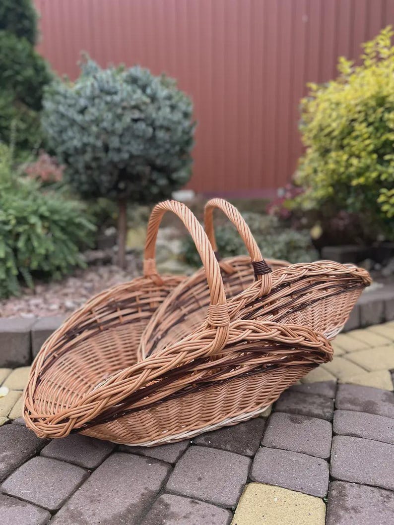 May include: A large, oval-shaped wicker basket featuring a woven light brown and dark brown pattern. The basket has two arched handles that meet in the center, a deep, open-top design, and a sturdy, braided rim construction made of natural willow.