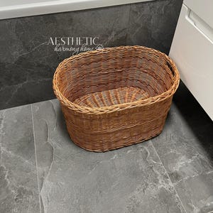 May include: A brown wicker oval laundry basket with a handle on the side. The basket is sitting on a gray tiled floor.