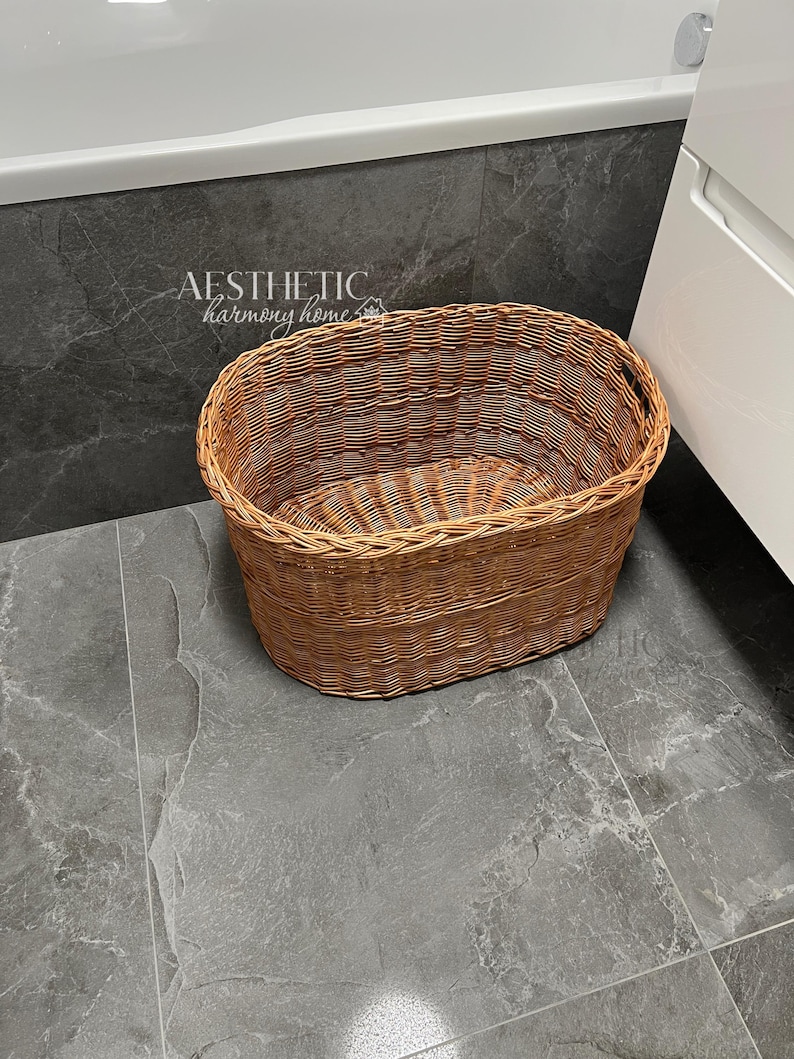 May include: A brown wicker oval laundry basket with a handle on the side. The basket is sitting on a gray tiled floor.