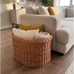 May include: A large, oval-shaped woven wicker basket with a natural light brown finish. The basket is filled with two rectangular throw pillows, one in mustard yellow and one in cream, along with a folded beige knit blanket tucked inside.