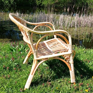 May include: A wicker chair with a light brown finish sits on a bed of green grass with small pink flowers. The chair has a rounded back and a woven seat.