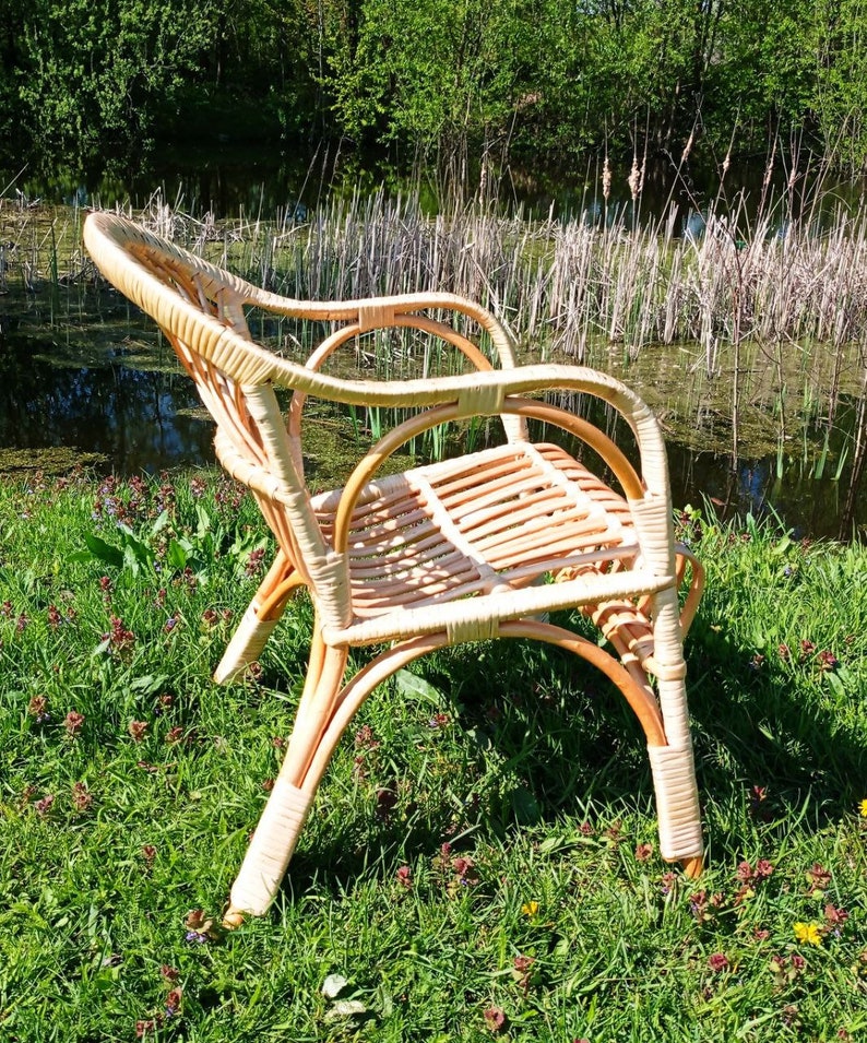May include: A wicker chair with a light brown finish sits on a bed of green grass with small pink flowers. The chair has a rounded back and a woven seat.
