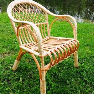 May include: A wicker chair with a light brown finish. The chair has a woven back and seat, and the arms are made of the same material. The chair is sitting on a patch of green grass.
