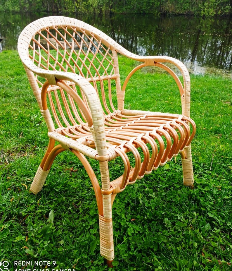 May include: A wicker chair with a light brown finish. The chair has a woven back and seat, and the arms are made of the same material. The chair is sitting on a patch of green grass.