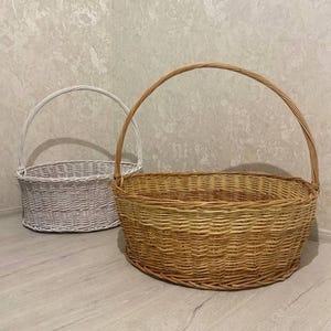 Op de afbeelding: Twee geweven rieten manden met gebogen handvatten. Eén mand is natuurlijk bruin, de andere is wit geschilderd. Beide manden zijn ovaal van vorm en staan op een lichtgekleurde houten vloer.