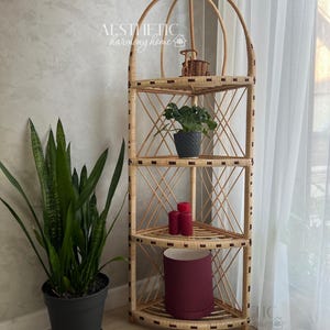 Puede incluir: Una estantería de esquina de tres niveles hecha de mimbre natural tejido con una parte superior redondeada. La estantería tiene un patrón de diamante y está decorada con una pequeña planta en maceta, velas y un objeto decorativo. La estantería está contra una pared blanca con una cortina blanca transparente en el fondo. El texto "AESTHETIC harmony home" es visible en la pared.