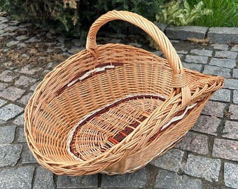 May include: A woven wicker basket with a curved handle. The basket is oval-shaped with a brown and white decorative pattern around the rim and base. The basket is made of light brown wicker and sits on a stone surface.