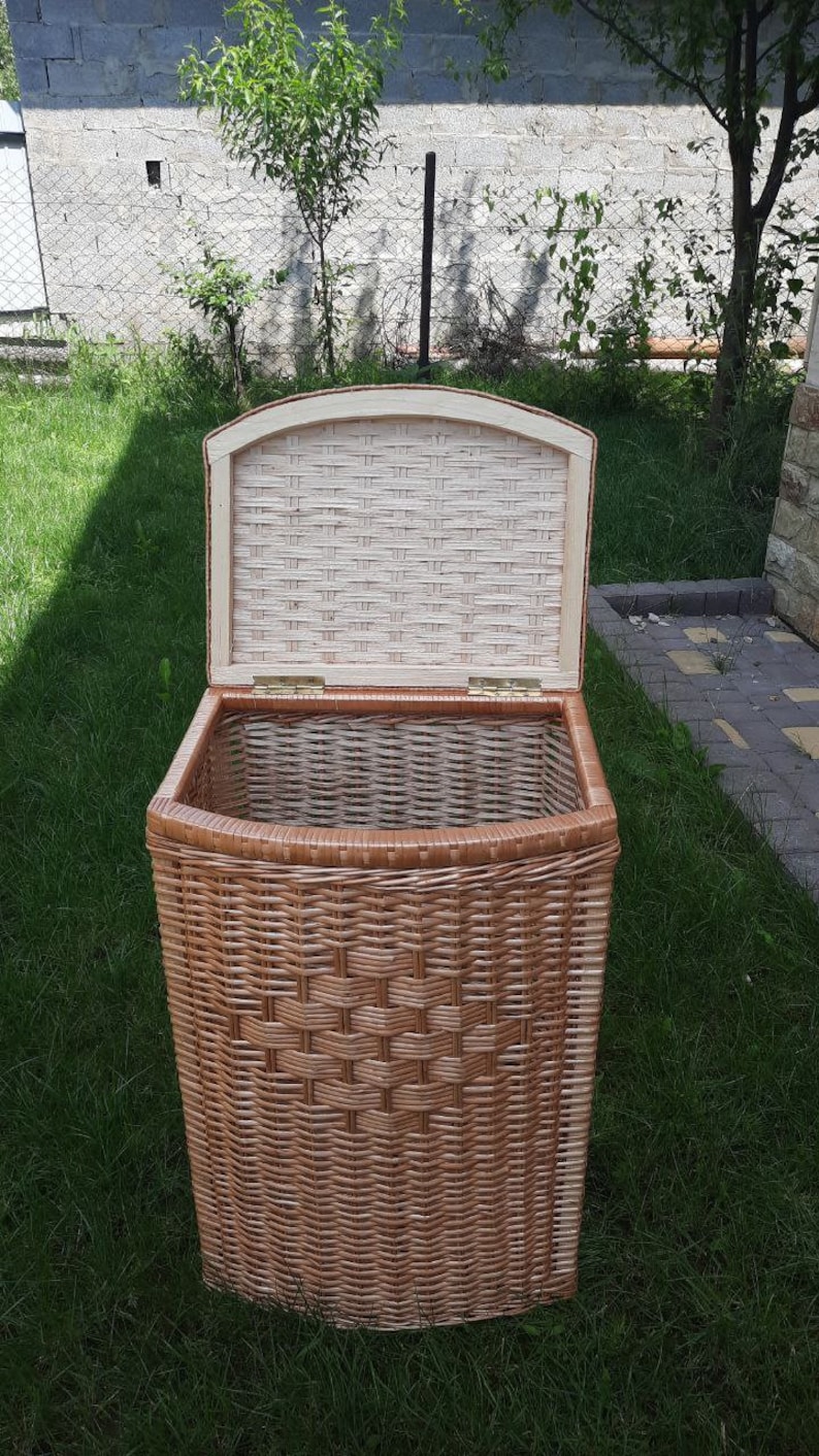 May include: A wicker storage basket with a lid, open and showing the inside of the basket. The basket is brown and has a woven pattern. The basket is sitting on a green lawn.