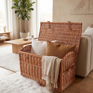 May include: A rectangular woven wicker storage trunk with an open hinged lid. The trunk features a natural light brown finish and contains a white pillow, a brown pillow, and a cream-colored throw blanket with fringe draped over the front edge.